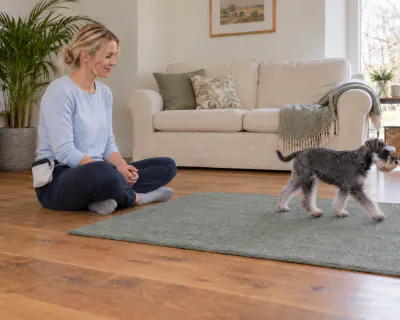 The puppy walking off the mat away from the woman, who stays sitting with her hands in her lap.